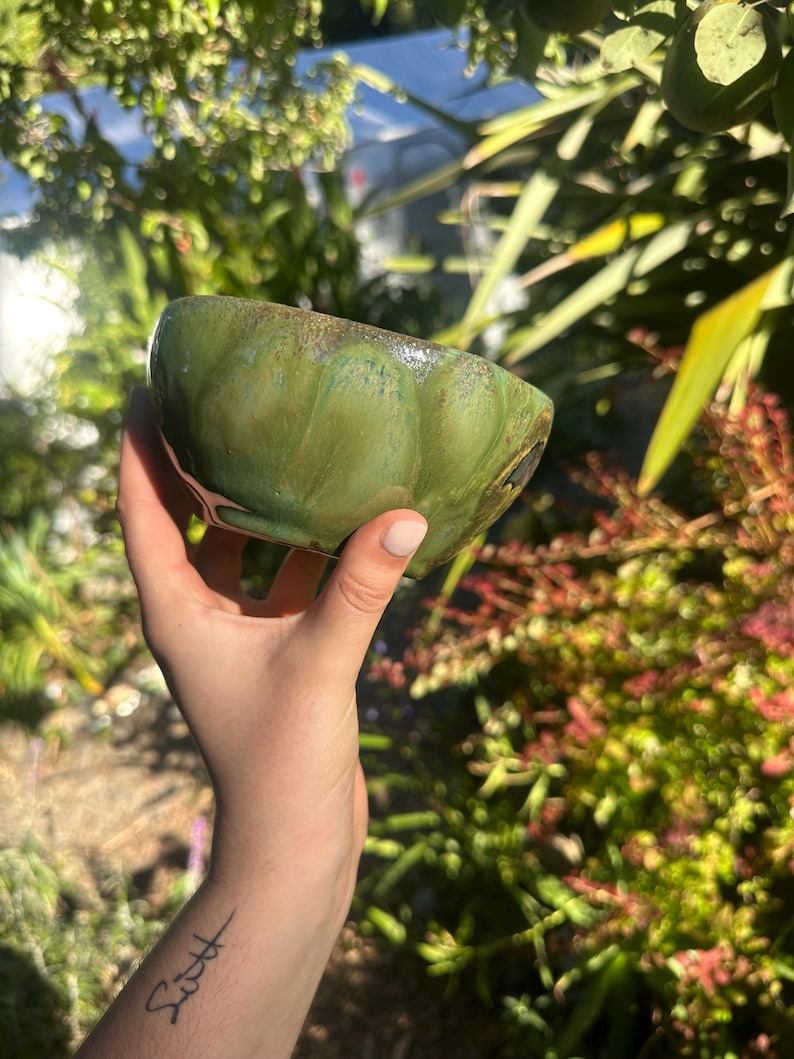 May include: A handmade, green ceramic bowl with a textured surface and a glossy finish. The bowl has a unique, organic shape and a dark brown rim. The bowl is held in a hand against a blurred background of green foliage.