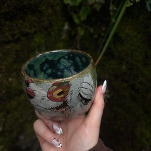 Puede incluir: Una peque&ntilde;a taza de cer&aacute;mica redonda con un exterior texturizado de color claro que presenta ilustraciones pintadas a mano de ojos estilizados y motivos circulares rojos. El interior de la taza est&aacute; esmaltado en un tono azul verdoso profundo.