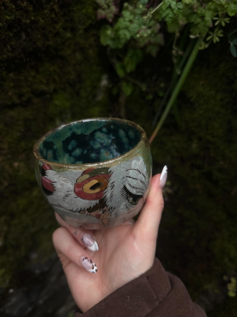 Puede incluir: Una peque&ntilde;a taza de cer&aacute;mica redonda con un exterior texturizado de color claro que presenta ilustraciones pintadas a mano de ojos estilizados y motivos circulares rojos. El interior de la taza est&aacute; esmaltado en un tono azul verdoso profundo.