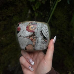 Puede incluir: Una peque&ntilde;a taza de cer&aacute;mica redonda con una ilustraci&oacute;n pintada a mano de un personaje de cabello casta&ntilde;o y una gran criatura blanca similar a un lobo con un ojo verde prominente. La taza tiene una base texturizada en tonos tierra.
