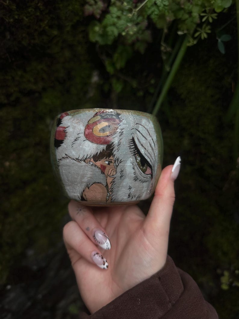 Puede incluir: Una peque&ntilde;a taza de cer&aacute;mica redonda con una ilustraci&oacute;n pintada a mano de un personaje de cabello casta&ntilde;o y una gran criatura blanca similar a un lobo con un ojo verde prominente. La taza tiene una base texturizada en tonos tierra.