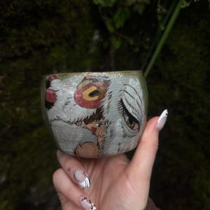 Puede incluir: Una taza de cer&aacute;mica con una ilustraci&oacute;n pintada a mano de un personaje de cabello casta&ntilde;o con una marca roja en la mejilla, junto a una gran criatura blanca similar a un lobo con un ojo verde y un detalle circular rojo en la cabeza.