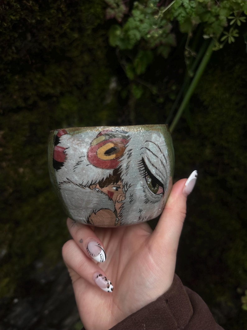 Puede incluir: Una taza de cer&aacute;mica con una ilustraci&oacute;n pintada a mano de un personaje de cabello casta&ntilde;o con una marca roja en la mejilla, junto a una gran criatura blanca similar a un lobo con un ojo verde y un detalle circular rojo en la cabeza.
