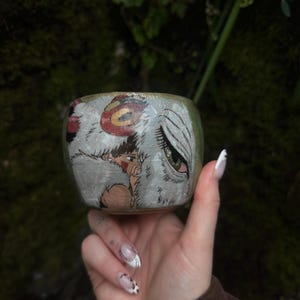 Puede incluir: Una peque&ntilde;a taza de cer&aacute;mica con una ilustraci&oacute;n pintada a mano de un personaje de cabello oscuro y un gran lobo blanco con un ojo verde. La obra est&aacute; realizada en tonos blancos, marrones y rojos sobre una superficie esmaltada clara.