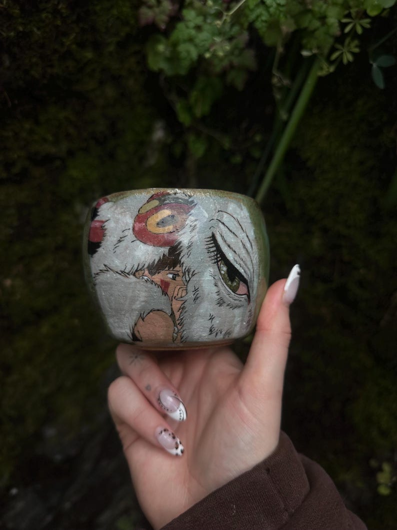 Puede incluir: Una peque&ntilde;a taza de cer&aacute;mica con una ilustraci&oacute;n pintada a mano de un personaje de cabello oscuro y un gran lobo blanco con un ojo verde. La obra est&aacute; realizada en tonos blancos, marrones y rojos sobre una superficie esmaltada clara.