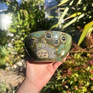 May include: A ceramic bowl with a hand-painted design featuring a cartoon character with an umbrella. The bowl has a green and brown glaze with black soot sprites. The bowl is held in a hand.