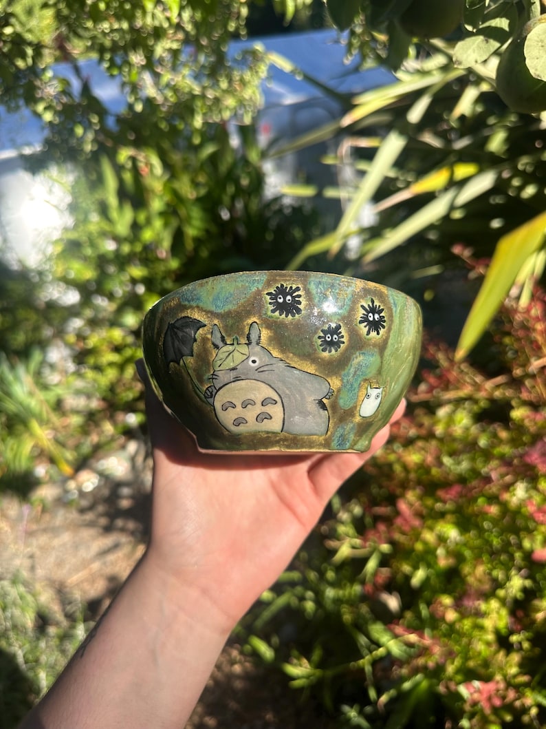 May include: A ceramic bowl with a hand-painted design featuring a cartoon character with an umbrella. The bowl has a green and brown glaze with black soot sprites. The bowl is held in a hand.