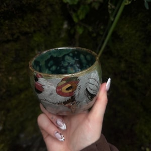 Puede incluir: Una peque&ntilde;a taza de cer&aacute;mica hecha a mano, de forma redondeada, con un interior verde oscuro moteado y brillante. El exterior presenta una ilustraci&oacute;n pintada a mano de un personaje con ojos grandes, un detalle circular rojo y texturas blancas.