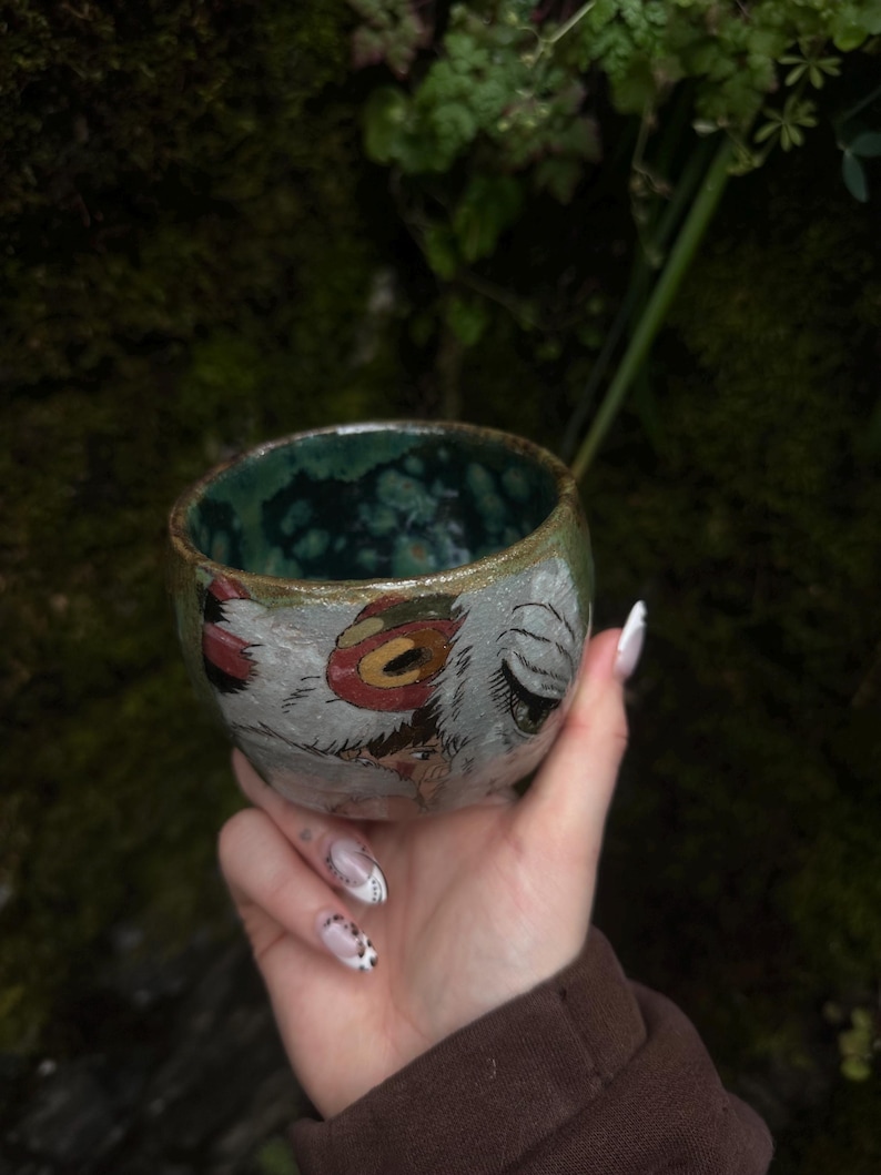 Puede incluir: Una peque&ntilde;a taza de cer&aacute;mica hecha a mano, de forma redondeada, con un interior verde oscuro moteado y brillante. El exterior presenta una ilustraci&oacute;n pintada a mano de un personaje con ojos grandes, un detalle circular rojo y texturas blancas.