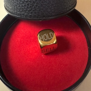K&ouml;nnte beinhalten: Ein goldfarbener W&uuml;rfel mit den eingravierten W&ouml;rtern HOLD und SELL auf seinen Seiten, der auf einer runden roten Samtunterlage in einem schwarzen zylindrischen Beh&auml;lter mit der Pr&auml;gung WE ARE TRADERS auf der Au&szlig;enseite liegt.