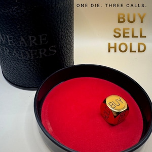K&ouml;nnte beinhalten: Ein schwarzer, texturierter zylindrischer Beh&auml;lter mit dem Text WE ARE TRADERS und ein passendes schwarzes Tablett mit rotem Filzinnenraum. Ein goldfarbener W&uuml;rfel liegt im Tablett mit den W&ouml;rtern BUY, SELL und HOLD. Text: ONE DIE. THREE CALLS. BUY SELL HOLD.
