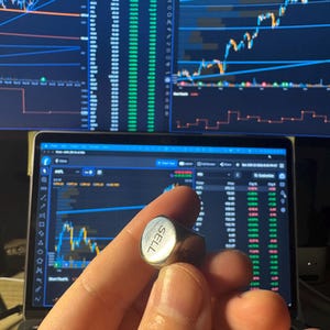 May include: A small, metallic, cube-shaped die with the word SELL engraved on one of its faces. The die is held in front of a laptop screen displaying financial stock market charts, data tables, and various technical analysis indicators.