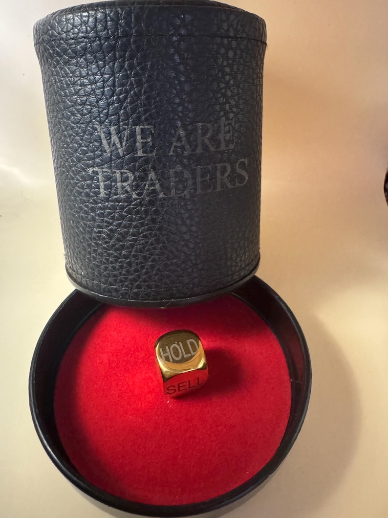 K&ouml;nnte beinhalten: Ein goldfarbener W&uuml;rfel mit den eingravierten W&ouml;rtern HOLD und SELL auf seinen Seiten, der auf einer runden roten Samtunterlage in einem schwarzen zylindrischen Beh&auml;lter mit der Pr&auml;gung WE ARE TRADERS auf der Au&szlig;enseite liegt.