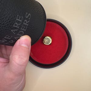 K&ouml;nnte beinhalten: Ein goldfarbener W&uuml;rfel mit dem eingravierten Wort BUY auf der Oberseite, der in einem runden schwarzen Beh&auml;lter mit rotem Samtinnenfutter liegt. Der schwarze Deckel tr&auml;gt den gepr&auml;gten Text WE ARE TRADERS auf der texturierten Au&szlig;enseite.