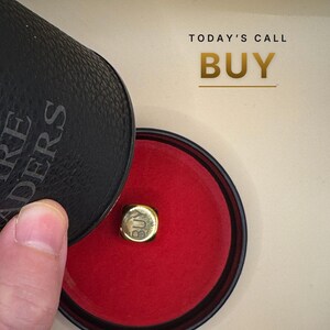 K&ouml;nnte beinhalten: Ein kleiner goldfarbener W&uuml;rfel mit dem eingravierten Wort BUY auf der Vorderseite, der in einem runden schwarzen Beh&auml;lter mit leuchtend rotem Innenfutter liegt. Der Text TODAY'S CALL BUY ist in Gold &uuml;ber dem Beh&auml;lter aufgedruckt.