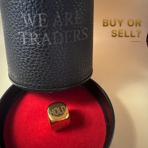 K&ouml;nnte beinhalten: Ein goldfarbener W&uuml;rfel mit den eingravierten W&ouml;rtern HOLD und SELL, der in einem schwarzen runden Beh&auml;lter mit rotem Innenraum liegt. Der Deckel des Beh&auml;lters tr&auml;gt den Text WE ARE TRADERS und BUY OR SELL?
