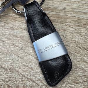 Könnte beinhalten: Schwarzer Leder-Schlüsselanhänger mit einem silbernen Metallband mit der Aufschrift "WE ARE TRADER". Der Schlüsselanhänger ist an einem silbernen Schlüsselring mit einem Schlüssel befestigt. Der Hintergrund ist eine hellbraune Holzoberfläche.