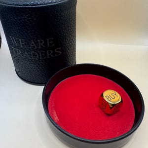 K&ouml;nnte beinhalten: Ein schwarzer zylindrischer Beh&auml;lter mit dem eingepr&auml;gten Text WE ARE TRADERS an der Seite, kombiniert mit einer schwarzen runden Schale mit leuchtend rotem Samtfutter und einem einzelnen goldenen W&uuml;rfel mit den W&ouml;rtern BUY und HOLD.