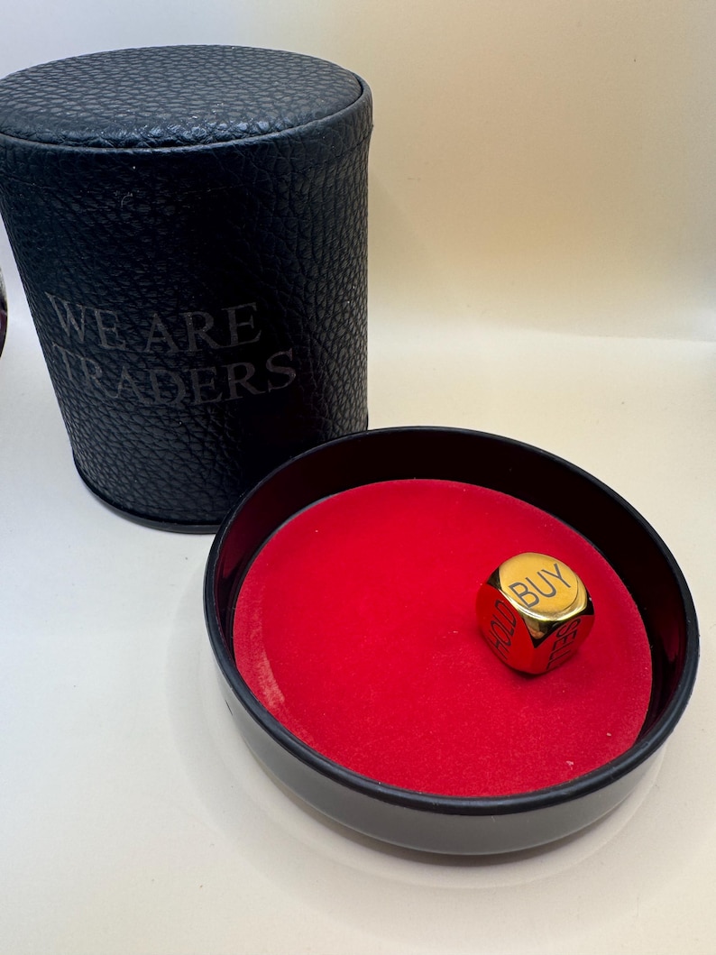 K&ouml;nnte beinhalten: Ein schwarzer zylindrischer Beh&auml;lter mit dem eingepr&auml;gten Text WE ARE TRADERS an der Seite, kombiniert mit einer schwarzen runden Schale mit leuchtend rotem Samtfutter und einem einzelnen goldenen W&uuml;rfel mit den W&ouml;rtern BUY und HOLD.