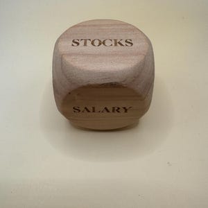 Może przedstawiać: Jasny drewniany sześcian z zaokrąglonymi krawędziami, z wygrawerowanym słowem STOCKS na górnej ściance oraz słowem SALARY na przedniej ściance. Słoje drewna są widoczne na całej powierzchni sześcianu, który posiada gładkie wykończenie.