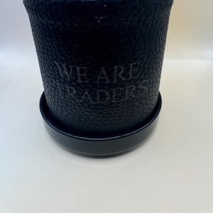 K&ouml;nnte beinhalten: Ein zylindrischer schwarzer Beh&auml;lter mit einer texturierten, genarbten Oberfl&auml;che und einem passenden Deckel. Auf der Vorderseite des Beh&auml;lters befindet sich der gepr&auml;gte Text "WE ARE TRADERS" in einer dunklen, dezenten Schrift.