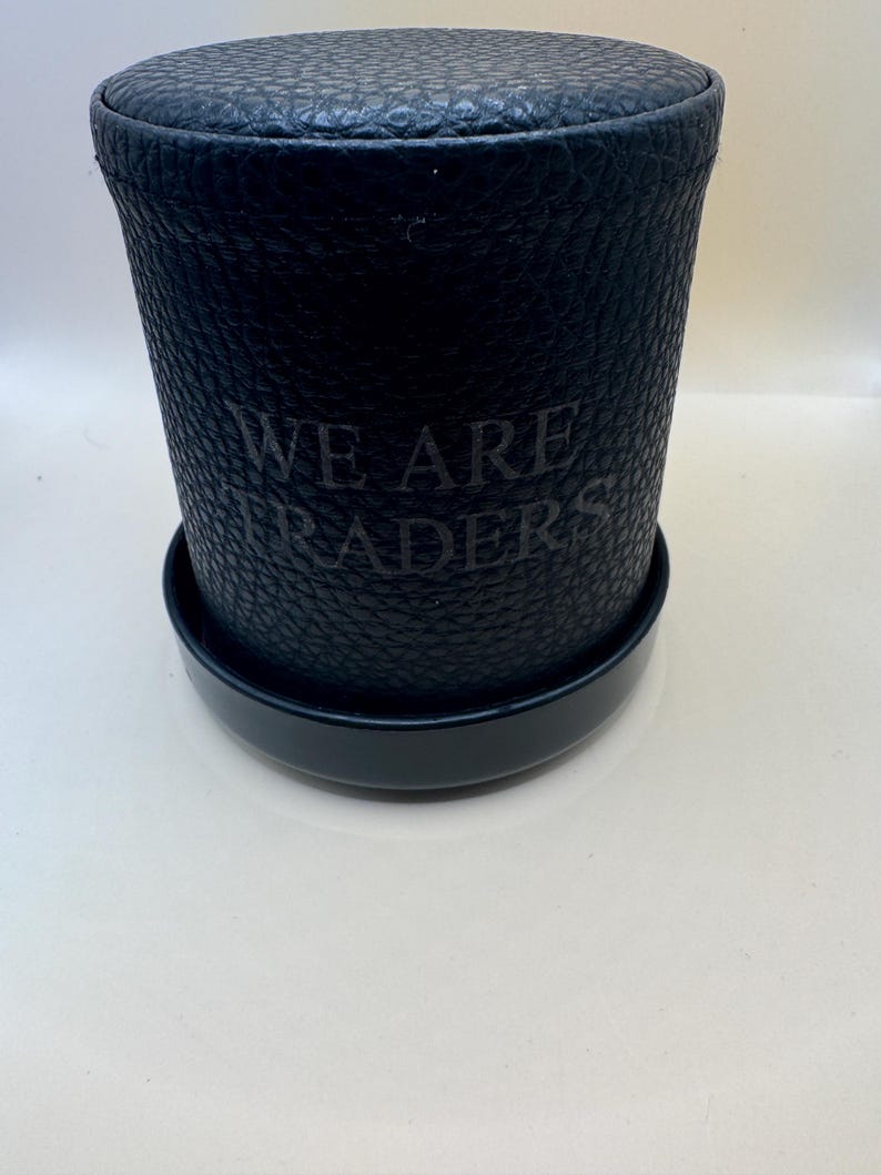 K&ouml;nnte beinhalten: Ein zylindrischer schwarzer Beh&auml;lter mit einer texturierten, genarbten Oberfl&auml;che und einem passenden Deckel. Auf der Vorderseite des Beh&auml;lters befindet sich der gepr&auml;gte Text "WE ARE TRADERS" in einer dunklen, dezenten Schrift.