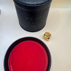 K&ouml;nnte beinhalten: Ein schwarzer zylindrischer Beh&auml;lter mit strukturierter Oberfl&auml;che und dem gepr&auml;gten Text WE ARE TRADERS, dazu ein separater schwarzer Deckel mit leuchtend rotem Innenfutter und ein goldfarbener W&uuml;rfel mit dem Wort BUY.