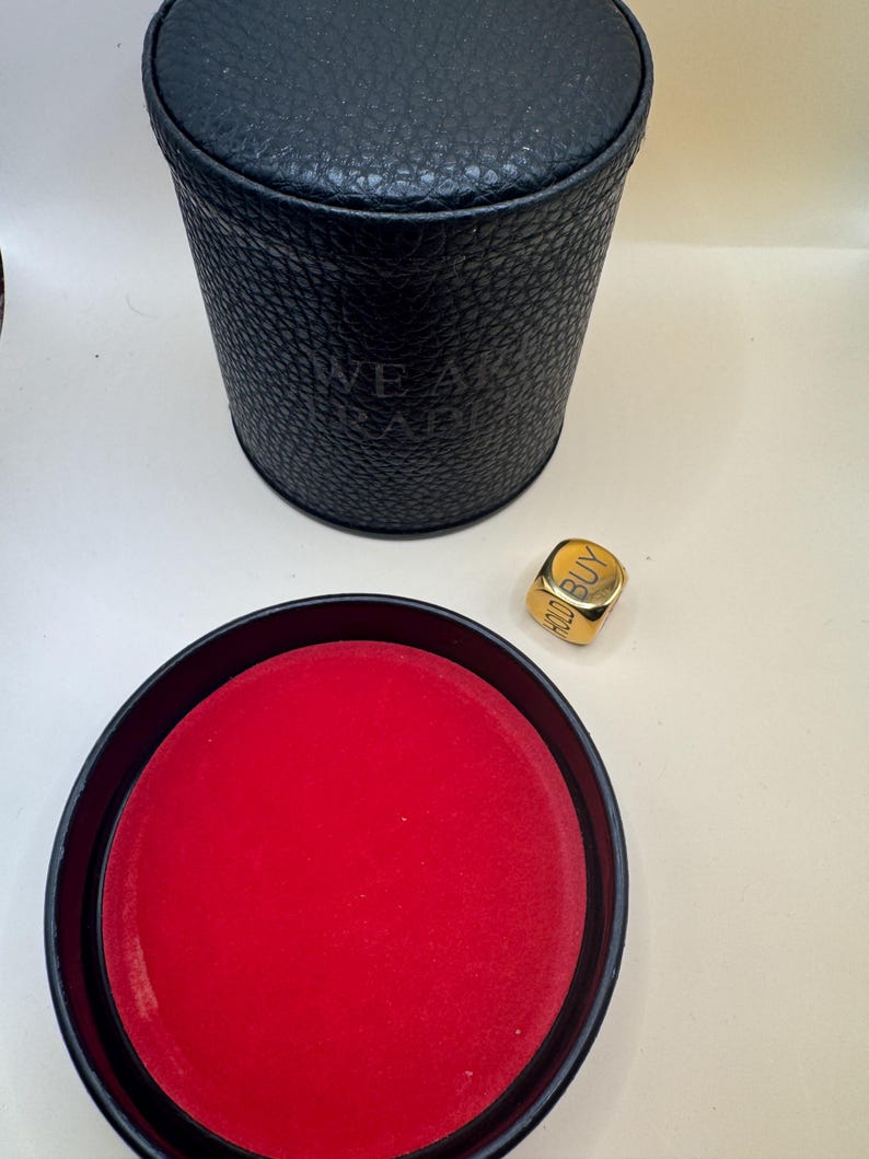 K&ouml;nnte beinhalten: Ein schwarzer zylindrischer Beh&auml;lter mit strukturierter Oberfl&auml;che und dem gepr&auml;gten Text WE ARE TRADERS, dazu ein separater schwarzer Deckel mit leuchtend rotem Innenfutter und ein goldfarbener W&uuml;rfel mit dem Wort BUY.