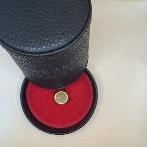 K&ouml;nnte beinhalten: Ein schwarzes zylindrisches Etui mit strukturierter Oberfl&auml;che und dem eingepr&auml;gten Text WE ARE TRADER an der Seite. Der Deckel ist ge&ouml;ffnet und gibt den Blick auf ein leuchtend rotes Innenfutter mit einem goldenen quadratischen Ring frei.