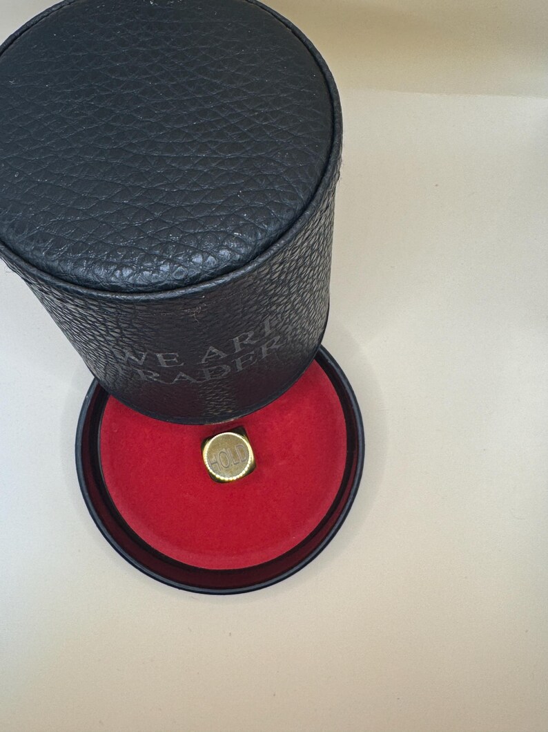 K&ouml;nnte beinhalten: Ein schwarzes zylindrisches Etui mit strukturierter Oberfl&auml;che und dem eingepr&auml;gten Text WE ARE TRADER an der Seite. Der Deckel ist ge&ouml;ffnet und gibt den Blick auf ein leuchtend rotes Innenfutter mit einem goldenen quadratischen Ring frei.