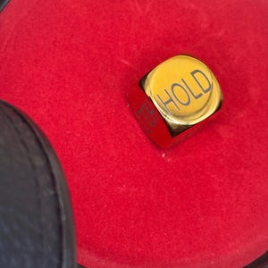 K&ouml;nnte beinhalten: Ein goldfarbener Metallw&uuml;rfel mit abgerundeten Kanten, auf dessen Oberseite das Wort HOLD in Schwarz eingraviert ist. Der W&uuml;rfel liegt in einem runden Beh&auml;lter, der mit leuchtend rotem Stoff ausgekleidet ist und einen schwarzen Rand hat.