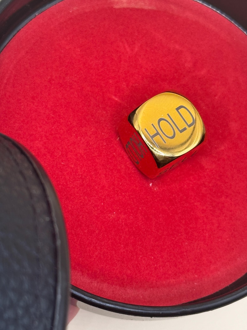 K&ouml;nnte beinhalten: Ein goldfarbener Metallw&uuml;rfel mit abgerundeten Kanten, auf dessen Oberseite das Wort HOLD in Schwarz eingraviert ist. Der W&uuml;rfel liegt in einem runden Beh&auml;lter, der mit leuchtend rotem Stoff ausgekleidet ist und einen schwarzen Rand hat.
