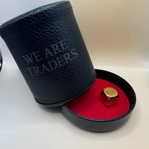 Può includere: Un contenitore cilindrico nero con superficie testurizzata e la scritta WE ARE TRADERS stampata sul lato. Il contenitore è aperto e rivela una base interna rosso vivo con un singolo dado cubico color oro che riporta la parola BUY in alto.