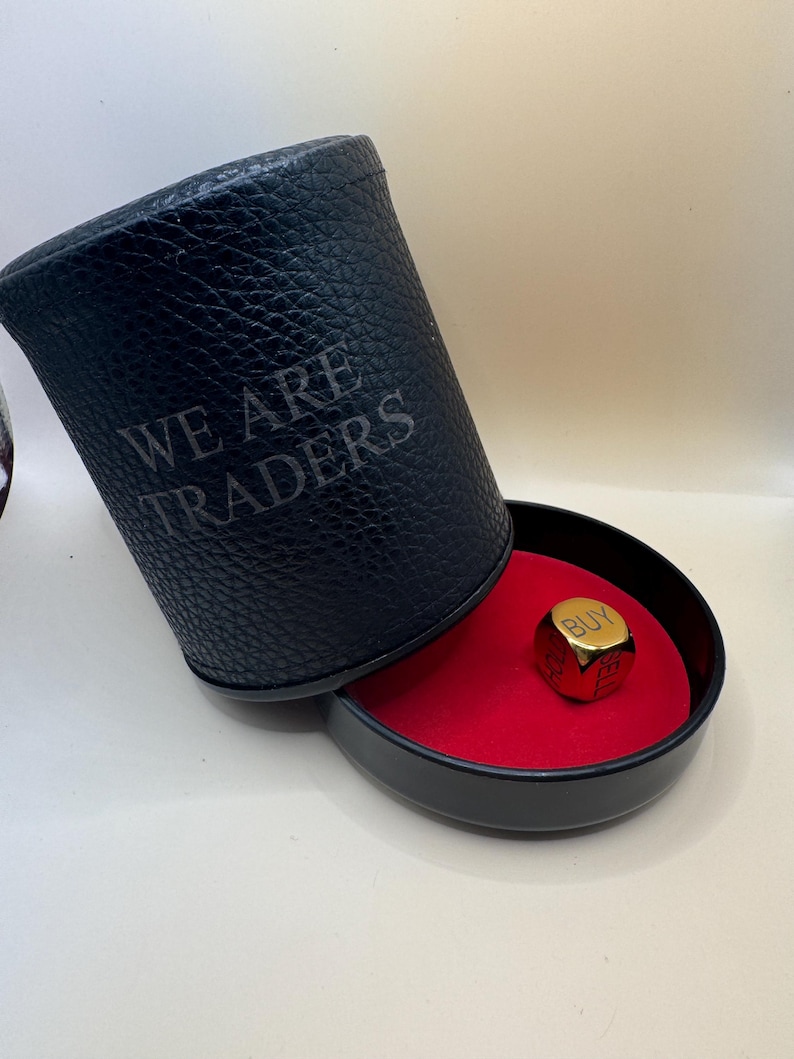K&ouml;nnte beinhalten: Ein schwarzer, zylindrischer W&uuml;rfelbecher in Lederoptik mit dem eingepr&auml;gten Text WE ARE TRADERS an der Seite. Der Becher ist geneigt und gibt den Blick auf einen goldfarbenen Metallw&uuml;rfel frei, dessen Oberseite das Wort BUY zeigt.