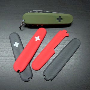 Op de afbeelding: Een olijfgroen Zwitsers zakmes met een zwart kruisembleem en verschillende vervangende schalen in rood en grijs. De schalen hebben een wit kruisontwerp. Het mes is ongeveer 9 cm lang.