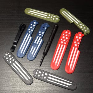 Puede incluir: Una colección de fundas para cuchillos y herramientas de varios colores, incluyendo fundas rojas, azules, verdes y grises. Cada funda presenta un diseño de estrellas y rayas blancas. También hay un clip de bolsillo negro y una herramienta negra.