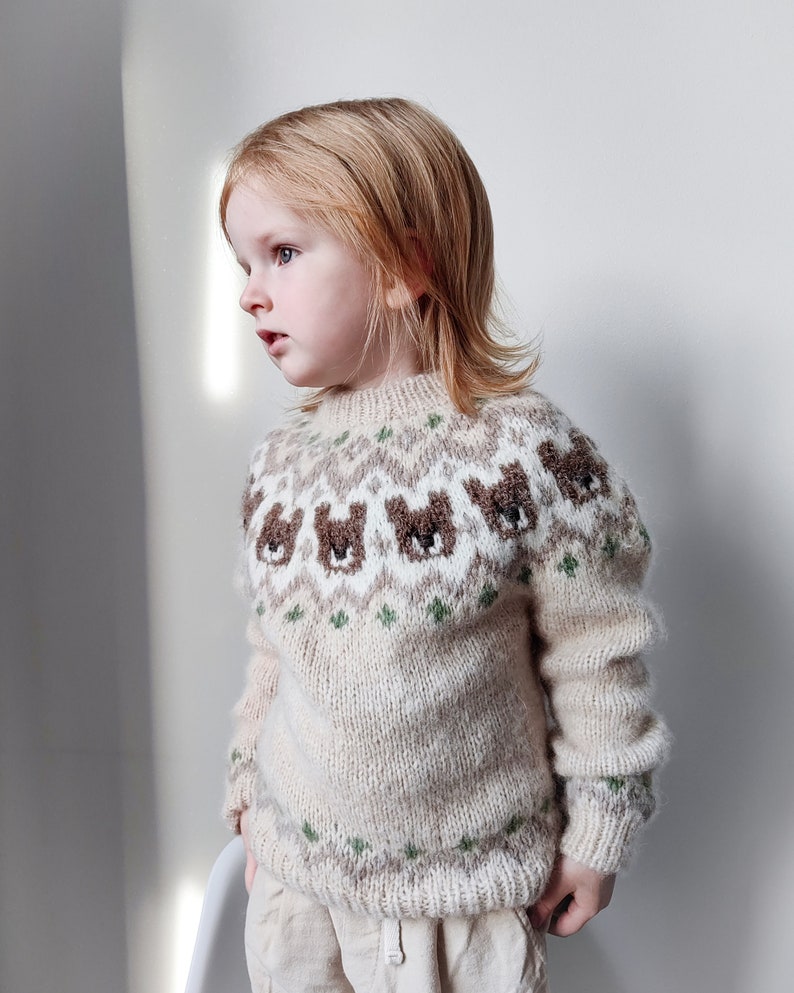 May include: A young child wearing a cream-colored knitted sweater with a brown bear pattern. The sweater has a green and white geometric design around the neckline.