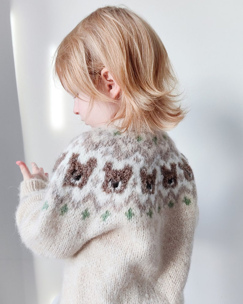 May include: A cream-colored knitted sweater with a brown bear pattern. The sweater has a crew neck and long sleeves.