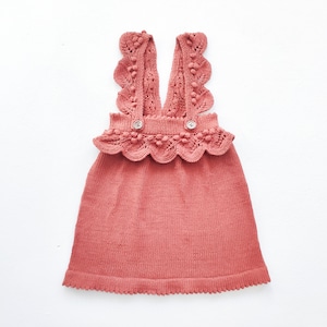 May include: A terracotta-colored knitted dress with a scalloped lace trim and pom-pom accents. The dress has a sleeveless design and a V-neckline.
