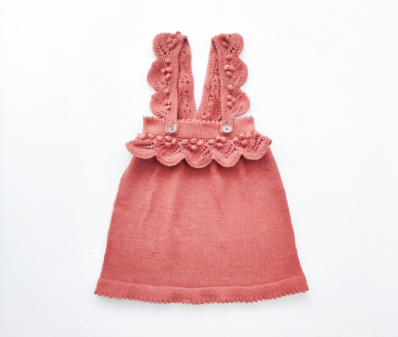 May include: A terracotta-colored knitted dress with a scalloped lace trim and pom-pom accents. The dress has a sleeveless design and a V-neckline.