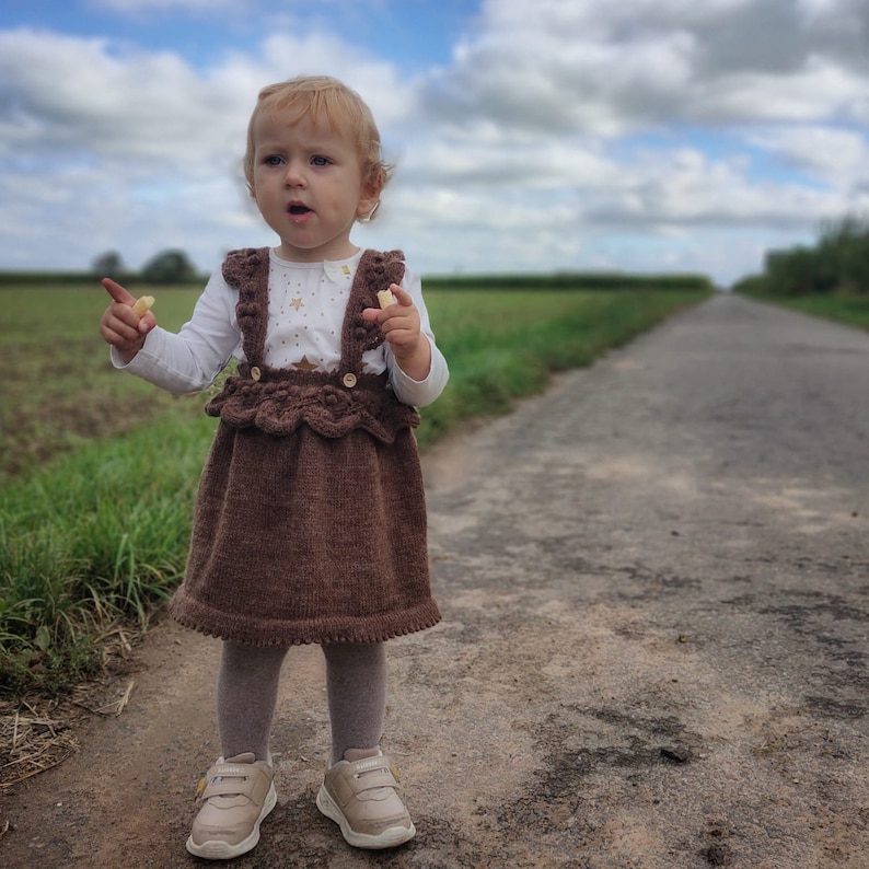 May include: A young child wearing a brown knitted dress with a white top and a brown knitted headband. The child is standing on a dirt road with green grass on either side.