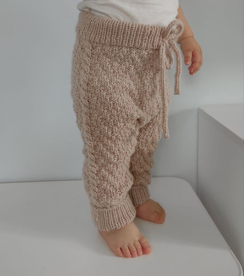 May include: Beige knitted baby trousers with a drawstring waist. The trousers have a cable knit pattern and are made of a soft, comfortable material.