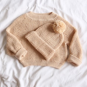 Puede incluir: Un su&eacute;ter de punto beige con un gorro de punto beige a juego con un pomp&oacute;n esponjoso.