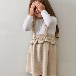 May include: A young girl wearing a white long-sleeved shirt and a beige knitted dress with a scalloped hem and wooden buttons. She is covering her eyes with her hands and smiling.