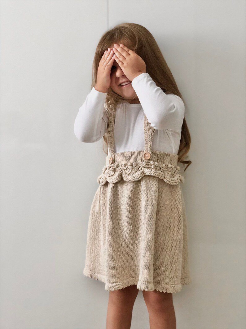 May include: A young girl wearing a white long-sleeved shirt and a beige knitted dress with a scalloped hem and wooden buttons. She is covering her eyes with her hands and smiling.
