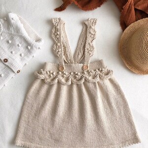 May include: A beige knitted dress with a scalloped hem and straps featuring crocheted details and small white pom-poms.