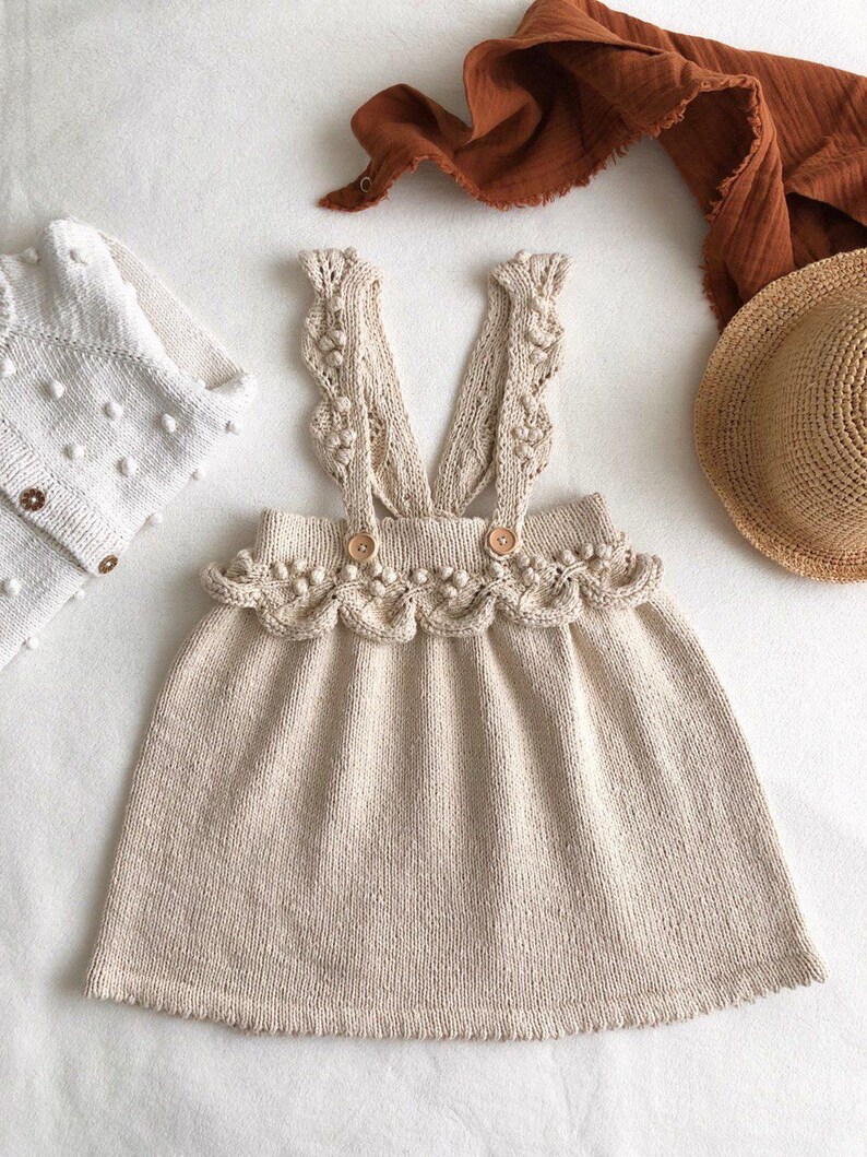 May include: A beige knitted dress with a scalloped hem and straps featuring crocheted details and small white pom-poms.