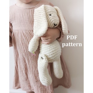 Op de afbeelding: Een wit gebreid konijn speelgoed met lange oren. Het konijn wordt vastgehouden door een kind dat een roze jurk draagt. De tekst "PDF pattern" is zichtbaar in de afbeelding.