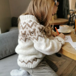 May include: A young person wearing a white and brown knitted sweater with a geometric pattern sits at a table and draws on a piece of paper with a red marker.