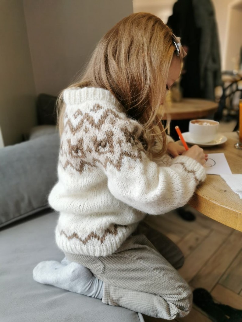 May include: A young person wearing a white and brown knitted sweater with a geometric pattern sits at a table and draws on a piece of paper with a red marker.